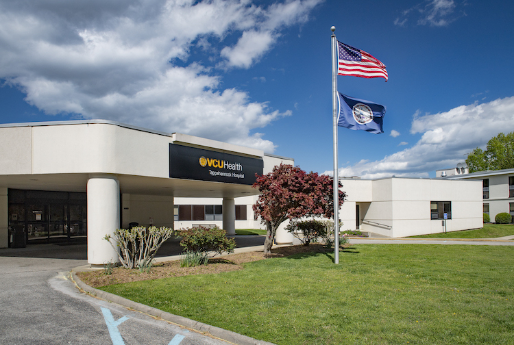 large white building with a black sign that says VCU Health Tappahannock Hospital. There is also a flag pole with an American flag and POW flag.