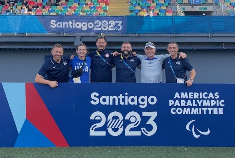 Group standing on a soccer field next to a sign for the Santiago 2023 games