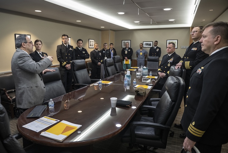 Group stands around a large table listening to a man speak.