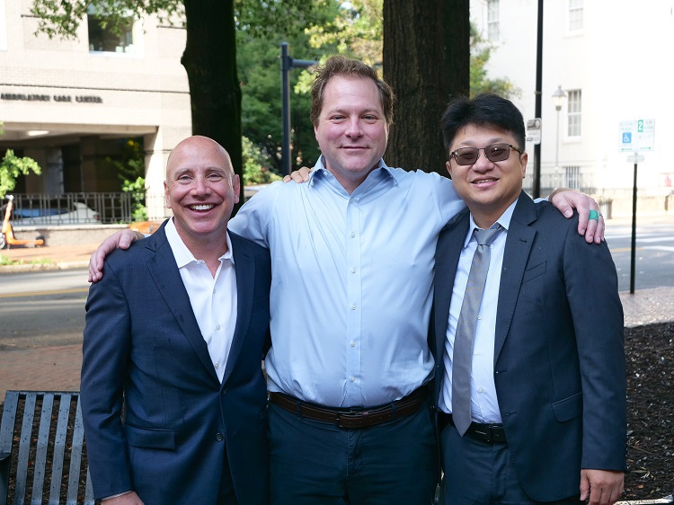 Team of doctors and surgeons stand together smiling outside of a hospital building.