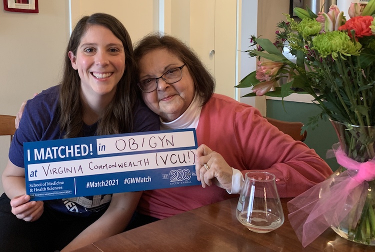 Daughter and mother hold up a sign smiling, the sign has information about where the daughter matched for her medical residency.