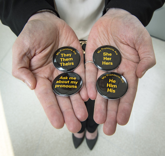 Four pins with a variety of pronouns on them lay flat on a person’s hands.