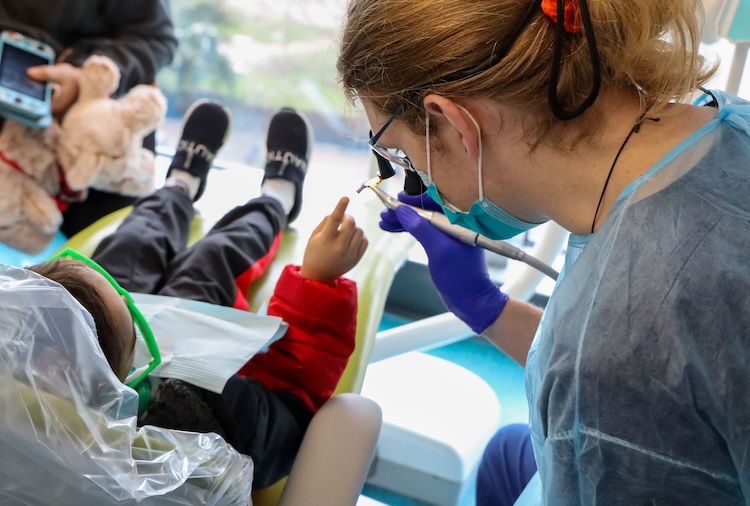 Small child sits in a chair and points to a device held by a dental care provider.