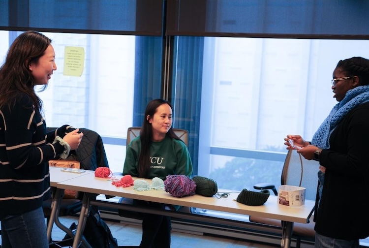 Three students talking to each other in a classroom. Some of them are holding yarn.