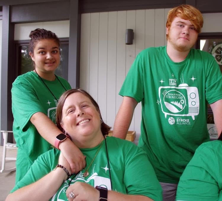 Mom sitting down holding her daughter’s hand. Son standing next to them smiling. They are all wearing green shirts.