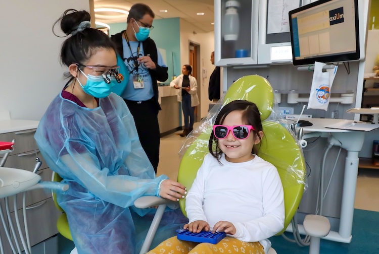 Little girl with sunglasses sits in a chair. Student stands next to her preparing for her appointment.