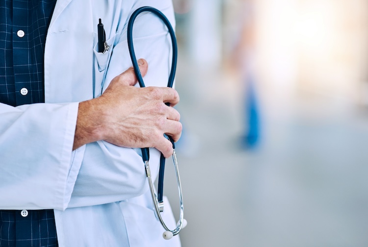 Doctor in a white coat stands with a stethoscope in hand and arms crossed. They are standing in a burry hallway and you cannot see their face. 