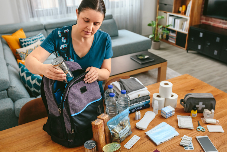 Woman putting cans of food to prepare emergency backpack in living room
