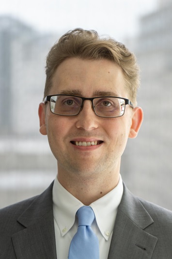 Man smiling for a headshot. He is wearing glasses and has a blue tie on with a grey suit.